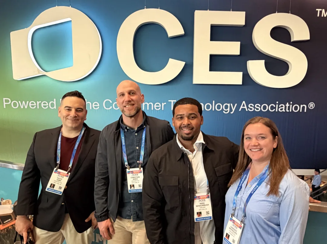 Four attendees wearing CES badges pose in front of a large CES logo